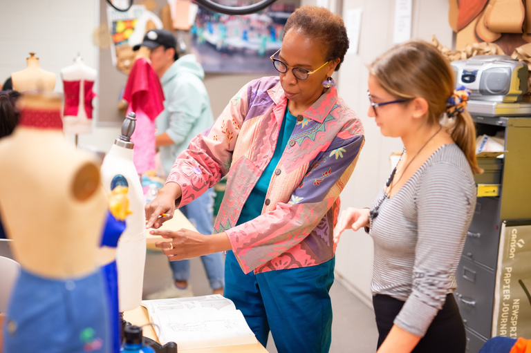 Loyce Arthur and student in Costume Shop working on a design. Loyce is wearing a pick blazer-type jacket with blue pants and the student had a black and white striped shirt with black pants. 
