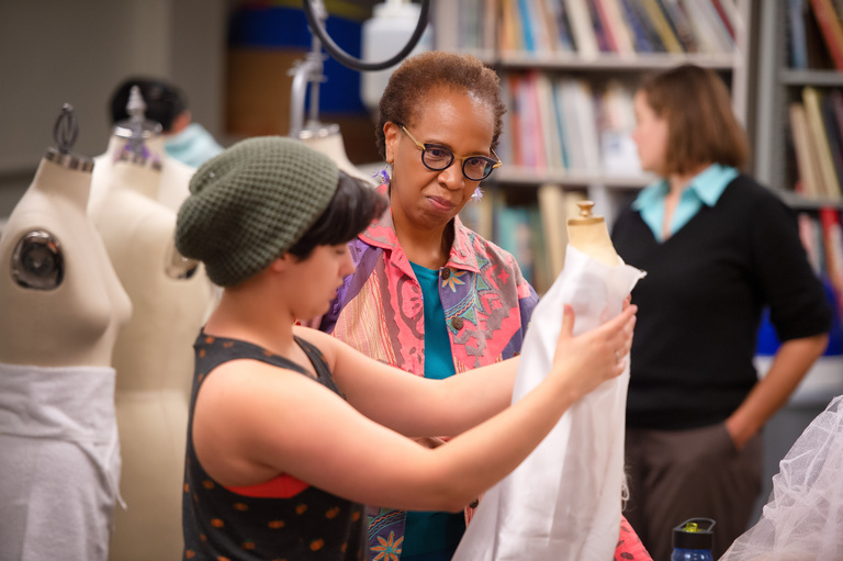Loyce Arthur observes student in Costume Shop working on a mannequin wrapped in white fabric. Loyce is wearing a pick blazer-type jacket with blue pants and the student is in grey beanie and a tank top.