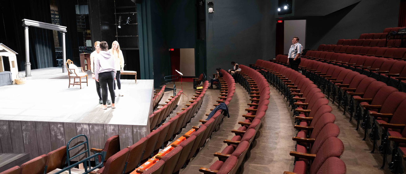 interior shot of Mabie Theatre showing student actors on the stage and a student director standing among the red audience seats, which are all empty