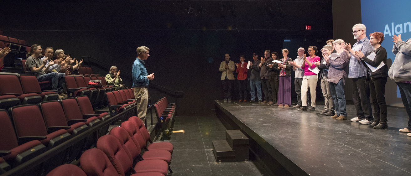 Interior shot of macvey theatre. Alan MacVey is standing in the audience, with Theatre faculty on the stage and in seats behind him applauding.