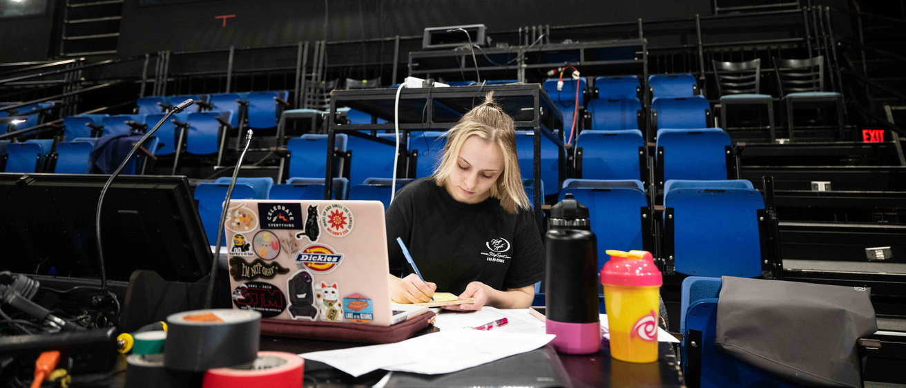 a female Theatre Design student with blonde hair sitting in the otherwise empty theatre working on her laptop with blue chairs and the sound booth behind her