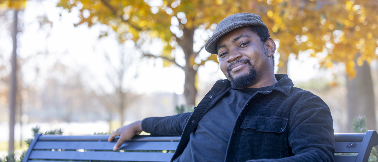Cheta Igbokwe sitting on a park bench outside the UI Theatre Building, with a tree in the background displaying golden fall colors