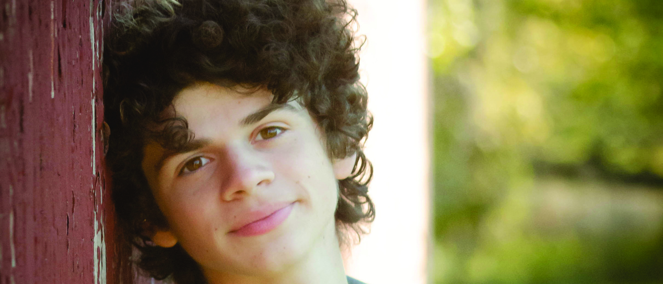 Jason Vernon, a second-year theatre and performing arts student leans against brick wall with yellow shirt and jean jacket