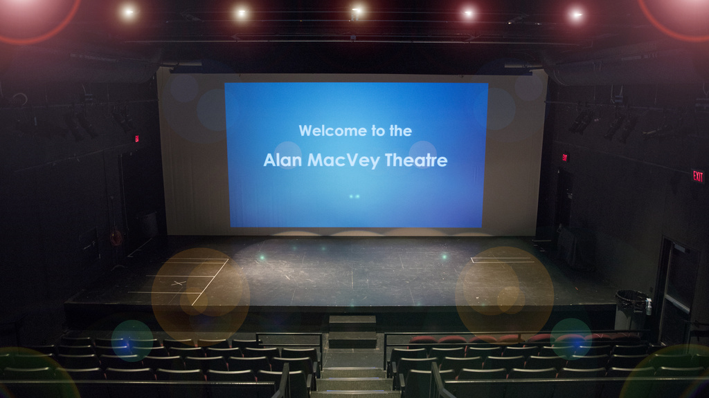 a view of the stage in MacVey Theatre from the audience's perspective. The stage and the seats are empty. On a screen onstage the words "welcome to MacVey Theatre" are projected in white on a blue background.